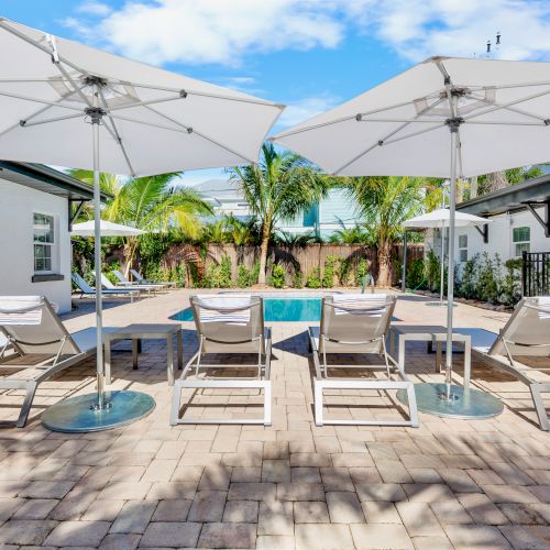 A poolside area features lounge chairs and large umbrellas under a clear blue sky, surrounded by palm trees and buildings.