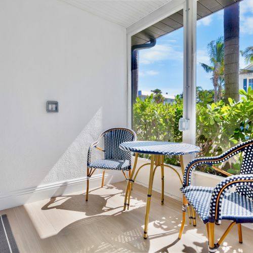 The image shows a cozy patio with two patterned chairs and a round table, overlooking lush greenery and palm trees under a bright blue sky.