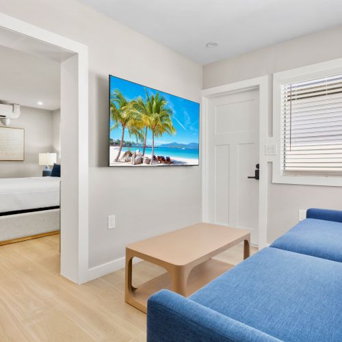 The image shows a small bright room with a blue sofa, wooden table, and wall art of a beach; a bedroom is visible through an open doorway.