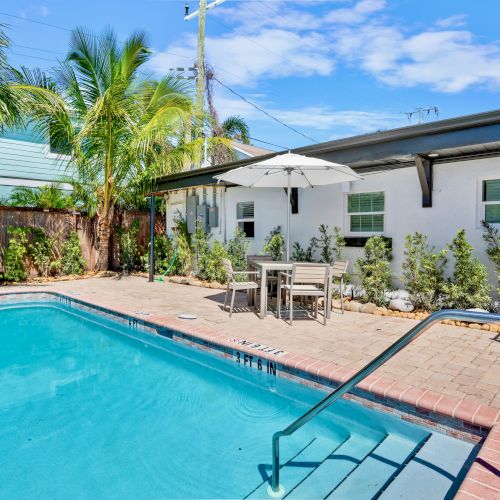 A backyard with a swimming pool, surrounded by palm trees and a white building. There's a patio area with a table and umbrella.
