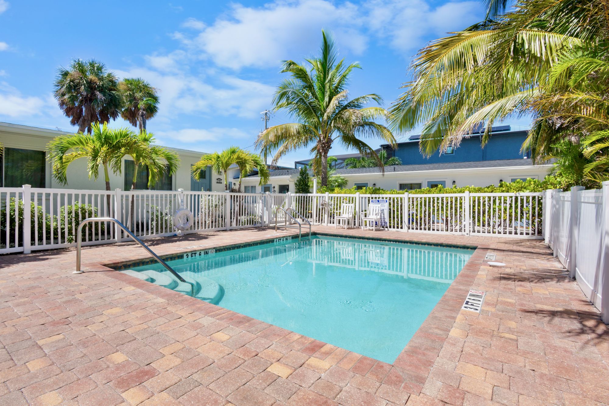 A small outdoor swimming pool is surrounded by a white fence and palm trees, with clear blue skies above.
