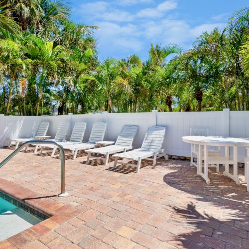 A poolside area with lounge chairs, a table set, surrounded by a fence and tropical trees, under a clear blue sky.