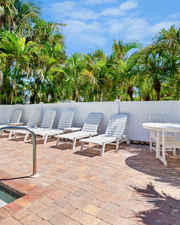 A poolside area with lounge chairs, a table set, surrounded by a fence and tropical trees, under a clear blue sky.