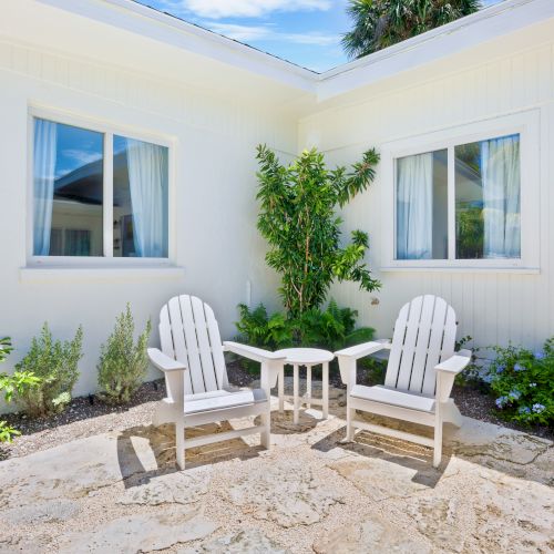Two white chairs with a table between them are set in a sunny outdoor patio area, surrounded by plants in front of a white building.