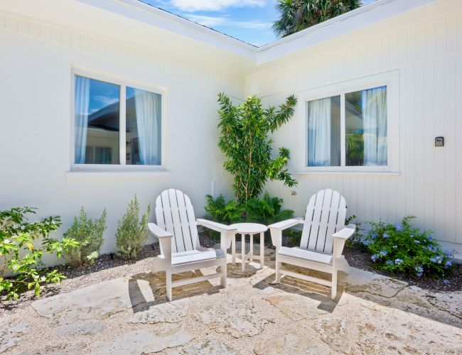 Two white chairs with a table between them are set in a sunny outdoor patio area, surrounded by plants in front of a white building.
