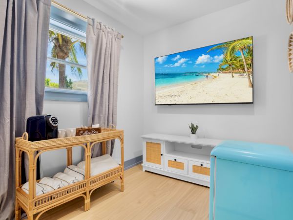 A room with a wicker console, a wall-mounted beach scene, a window with curtains, and a small turquoise ottoman.