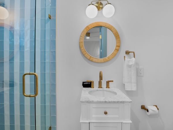 A bathroom with a round mirror, white vanity, gold fixtures, towel holder, and blue glass shower door is shown in the image.