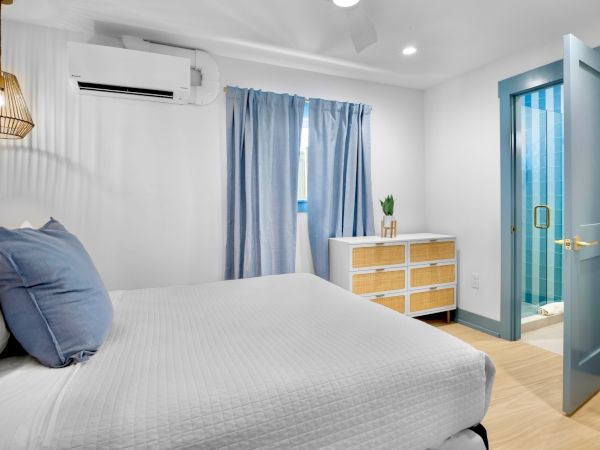 A cozy bedroom with a neatly made bed, blue curtains, a dresser, and an open door to a glass-walled shower in a brightly lit space.