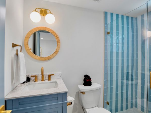 The image shows a bathroom with a blue vanity, round mirror, gold fixtures, and a shower with blue striped tiles.