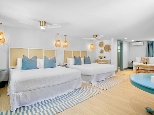 A bright hotel room with two large beds, blue accents, wicker lamps, and a cozy seating area, creating a welcoming and airy atmosphere.