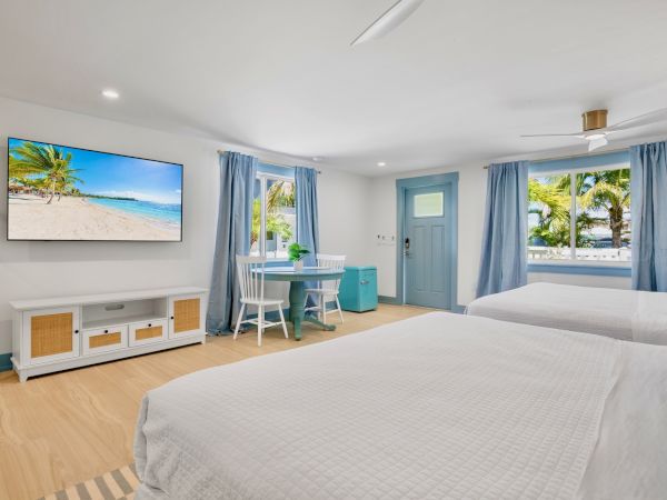 The image shows a bright, cozy room with two beds, a TV, and a small table by the window with blue curtains, offering a beach vibe.