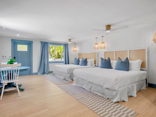 The image shows a bright, modern bedroom with two beds, blue accents, a desk with a chair, and pendant lighting, all on a wooden floor.