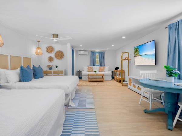 A bright, modern hotel room features two beds, a sitting area, wall art, and a large TV with blue accents and wooden flooring.
