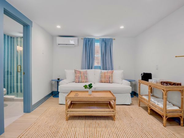 A cozy living room with a white sofa, wooden table, and blue curtains, featuring a wicker shelf and a light wooden floor.