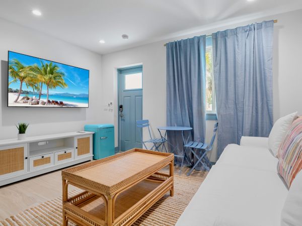 A cozy living room with a white couch, a wooden coffee table, a TV displaying a beach scene, a small dining set, and blue curtains.