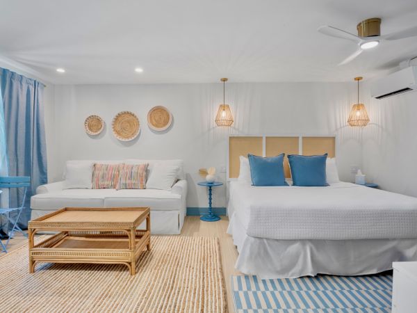 The image shows a bright, stylish bedroom with a white sofa, coffee table, bed with blue pillows, wicker pendant lights, and wall decor.