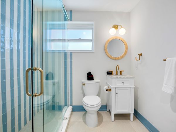 A bathroom with a glass shower, toilet, white vanity, and mirror. Blue-striped tiles are on the wall, with towels hanging nearby.
