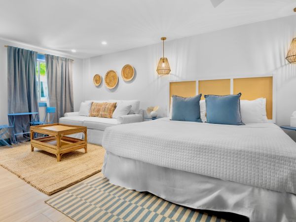 A cozy bedroom with a large bed, blue pillows, wicker lamps, a sofa, coffee table, and soft lighting, creating a warm, inviting ambiance.