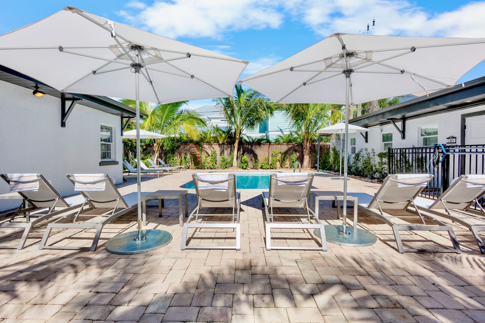 The image shows a poolside area with lounge chairs, umbrellas, palm trees, and a pool, all set under a clear blue sky.