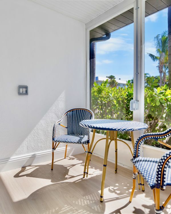 Two blue wicker chairs and a round table are on a sunlit patio with greenery and a view outside.