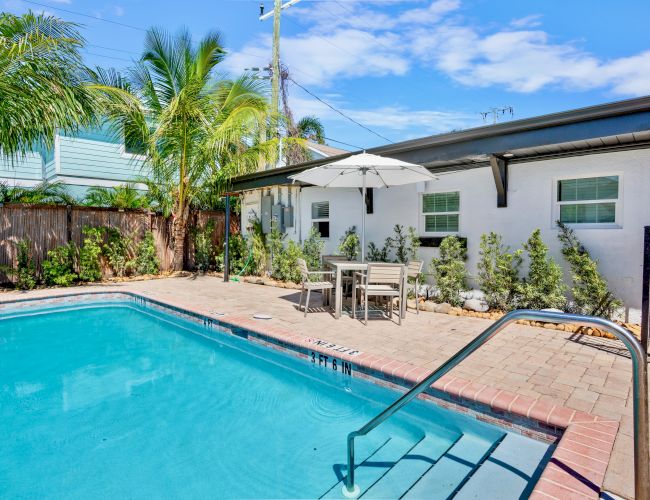 A backyard with a swimming pool, surrounded by palm trees and a patio area with a table and umbrella next to a building.