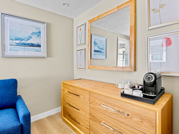 The image shows a cozy corner with a blue chair, a wooden dresser with a coffee maker, and various framed artworks on the walls.