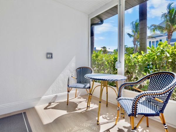 A sunlit patio features a small round table and two woven chairs with blue cushions, surrounded by lush greenery and palm trees outside.