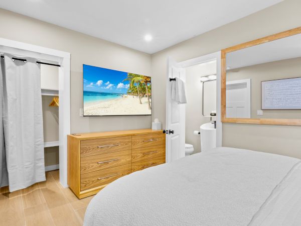 A bedroom with a bed, wooden dresser, wall-mounted beach scene TV, closet with curtain, and a door leading to a bathroom.