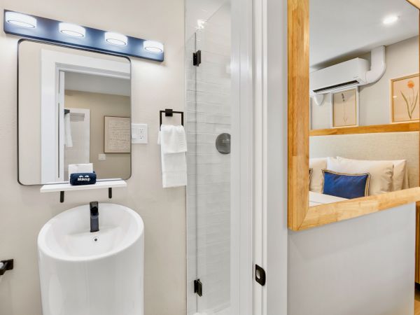 The image shows a modern bathroom with a sink, mirror, and nearby shower. There’s a glimpse into a bedroom area with a bed.