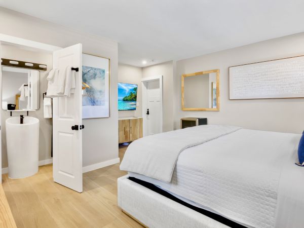 The image shows a modern bedroom with a bed, white linens, artwork on the walls, and a view into an en-suite bathroom with a sink and towels.