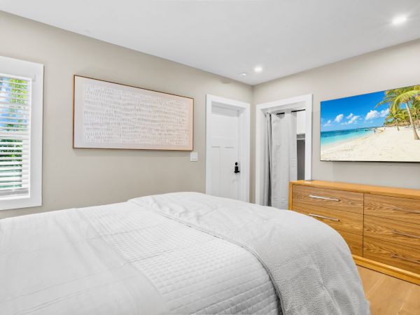 A bedroom with a bed, white linens, wooden dresser, abstract art, and a beach scene on the wall.