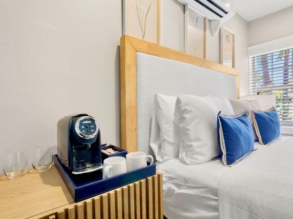A bedroom with a neatly made bed, blue pillows, and a side table holding a coffee maker and cups is shown here.