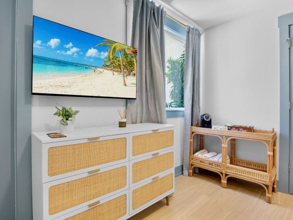 The image shows a room with a TV display of a beach scene, a white dresser with woven drawers, a rattan shelf, and a window with curtains.