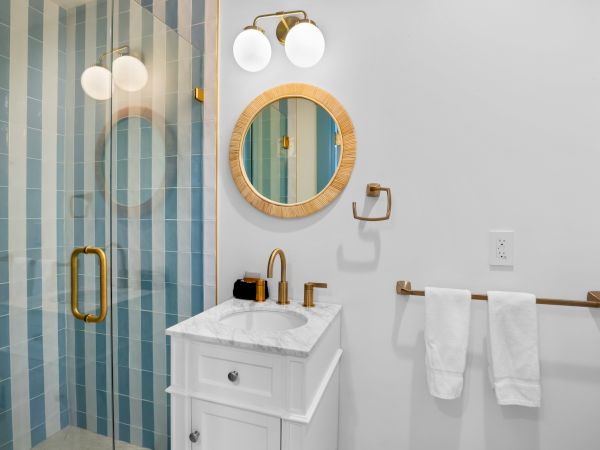 The image shows a bathroom with a white vanity, gold fixtures, round mirror, striped glass shower, and a towel rack with two towels.
