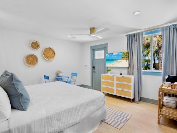 A bright bedroom with a bed, dresser, small table, and TV. There's a window and door with blue accents, and woven wall decorations.