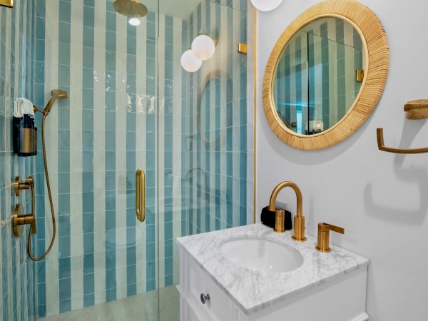 A bathroom with a round mirror, vanity, gold fixtures, and a shower with blue and white striped tiles is shown.