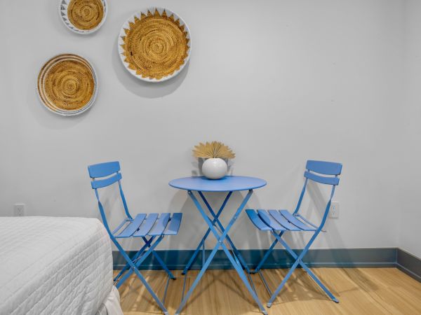 A small table with two blue chairs, a white vase on top, wall decor with woven patterns, and a segment of a bed; minimalist setting.