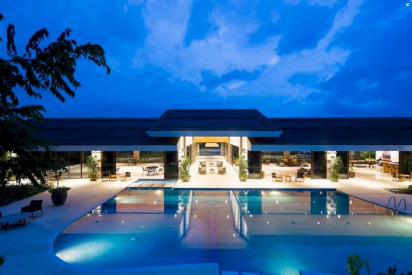A luxurious outdoor scene with a large illuminated pool, surrounded by modern architecture and seating areas under a twilight sky.