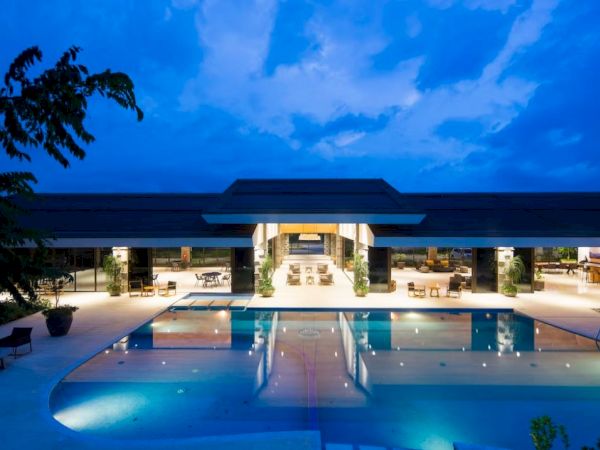 A luxurious outdoor scene with a large illuminated pool, surrounded by modern architecture and seating areas under a twilight sky.