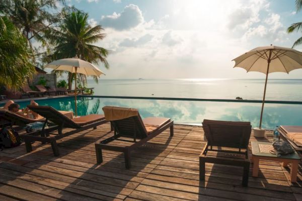 A tranquil poolside scene with lounge chairs, umbrellas, and palm trees against a sunny sky and calm water, creating a relaxing atmosphere.