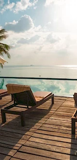 A tropical poolside scene with sun loungers, parasols, and palm trees overlooking a serene body of water under a cloudy sky.