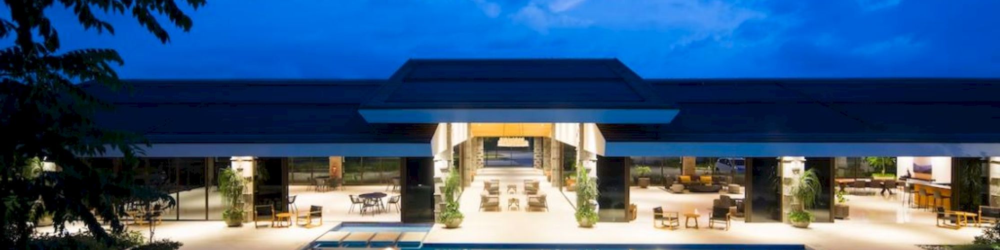 A luxurious outdoor area with a large pool, surrounded by a modern building and ambient evening lighting, under a partly cloudy sky.