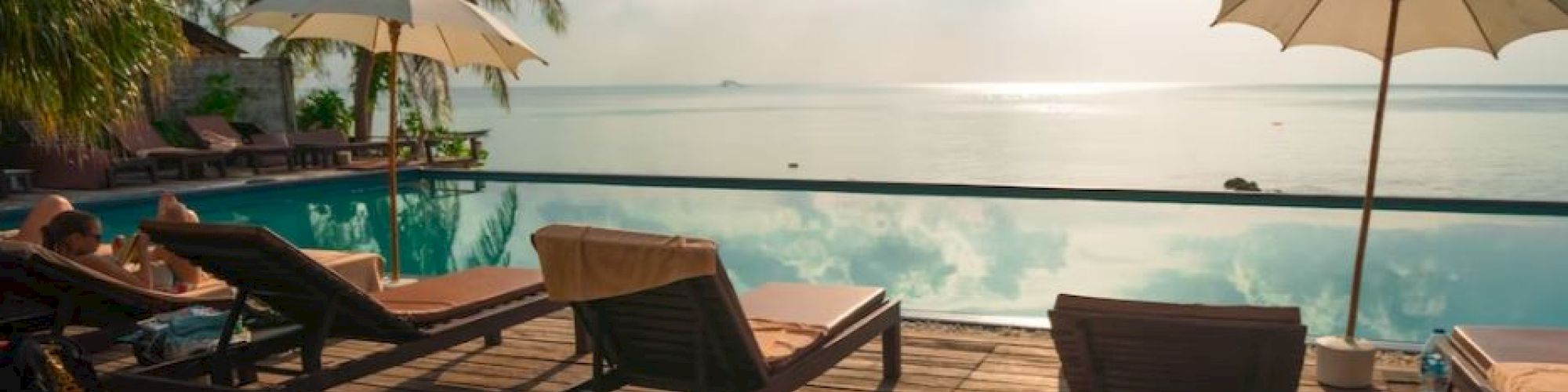 A serene poolside scene with lounge chairs, umbrellas, and palm trees overlooking the ocean under a cloudy sky.