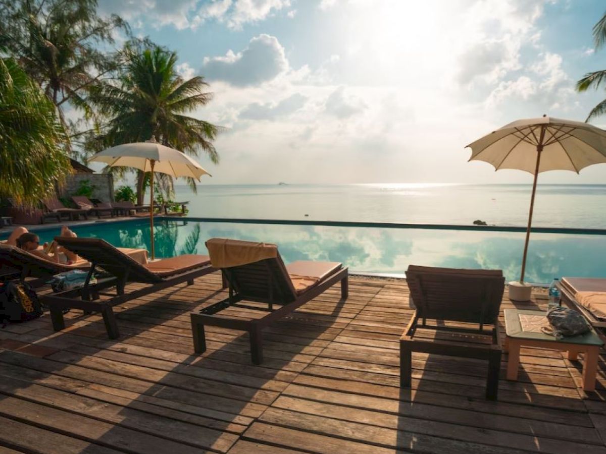 A serene poolside setting with lounge chairs, umbrellas, and palm trees overlooking a calm sea under a bright sky.