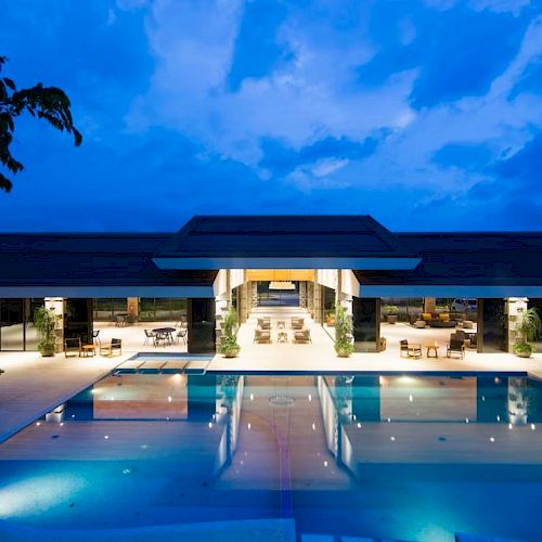 A luxurious building with a large illuminated pool, surrounded by a relaxing outdoor seating area under a vibrant blue evening sky.
