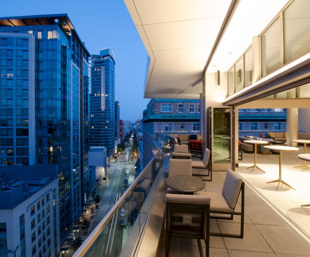 A modern urban balcony overlooking a cityscape with tall buildings, featuring outdoor seating and large windows in an evening setting.