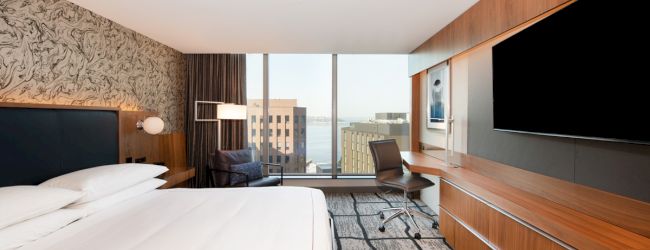 The image shows a modern hotel room with a large bed, TV, desk, and a window view of a cityscape and water.