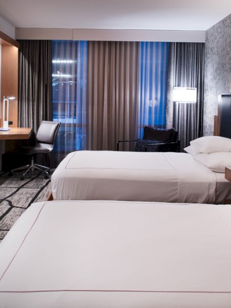 The image shows a modern hotel room with two beds, a desk, a TV, and stylish decor.