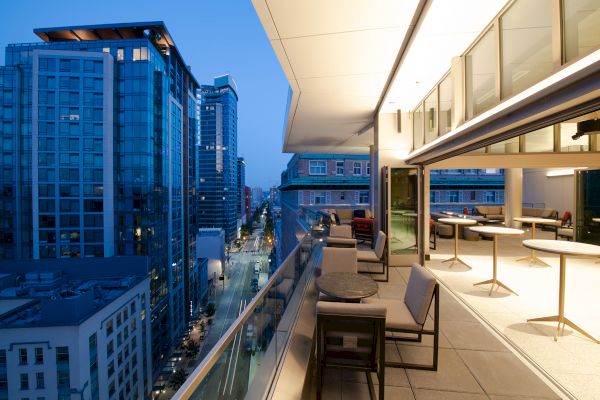 A modern balcony overlooks a cityscape at dusk with tall buildings and street view, featuring outdoor seating and warm lighting.