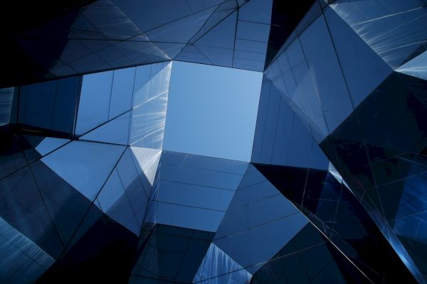 Looking up at geometric, glass-like structures converging towards a clear blue sky, forming a modern architectural pattern.